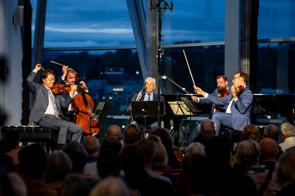 Musicians perform at dusk as the audience looks on, the river beyond the glass
